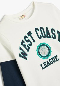 Biała i granatowa koszulka z długim rękawem z bawełny, z grafiką "WEST COAST LEAGUE" oraz emblematem koszykarskim w czarnych i turkusowych kolorach.