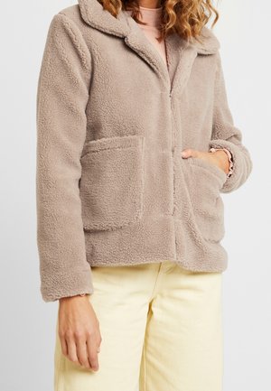 Mujer con chaqueta beige peluda con grandes bolsillos delanteros, camiseta rosa de manga larga y pantalones amarillo claro, mano en el bolsillo.