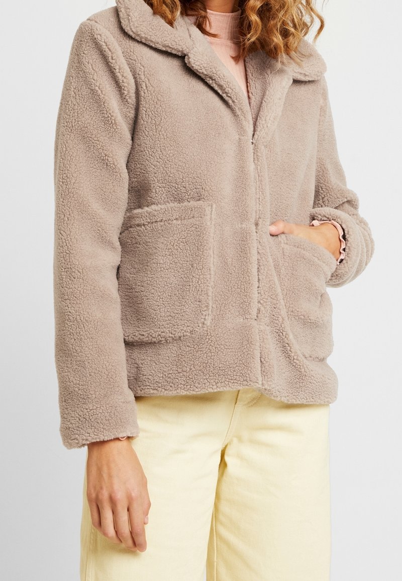Femme portant une veste beige duveteuse avec de grandes poches devant, un haut rose à manches longues et un pantalon jaune clair, main dans la poche.
