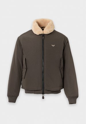 Homme portant une veste marron avec un col beige, une chemise bleu clair boutonnée, un jean bleu délavé et des bottes noires, debout avec une main dans la poche.