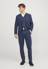 Costum trei piese bleu marin, realizat dintr-un material texturat, cu o croială ajustată și o vestă cu cinci nasturi. Cămașă albă și pantofi negri completează aspectul.