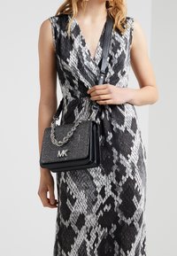 Borsa a tracolla a catena con motivo in bianco e nero, con un logo sulla parte anteriore e diverse tasche, tenuta da una persona.