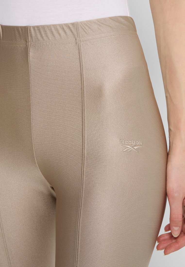Beżowe legginsy wykonane z gładkiego, strukturalnego materiału z elastycznym paskiem w talii oraz subtelnym haftowanym logo Reebok z boku.