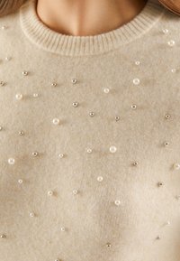 Pull en beige avec un col côtelé, orné de perles fantaisie et de perles argentées dispersées pour apporter texture et détails. Matière douce en tricot.