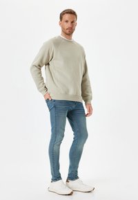 Felpa grigia chiara con scollatura rotonda e maniche raglan, abbinata a jeans blu attillati e sneakers bianche con dettagli marroni.