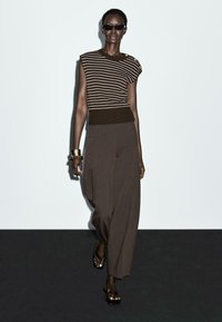Top a righe marrone in maglia con maniche corte e orlo a costine, abbinato a pantaloni marroni larghi. Gli accessori includono bracciali dorati e sandali neri.