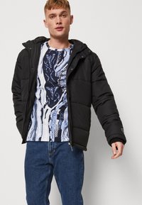 Svart polstret jakke med hette har glidelås og elastiske mansjetter, brukt over en blå og hvit mønstret skjorte og mørkeblå jeans.