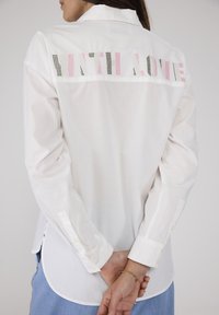 Weißes Hemd mit mehrfarbigem, gesticktem Text "MIT LIEBE" auf dem Rücken, langen Ärmeln und abgerundetem Saum.