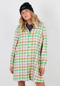Robe chemise à carreaux multicolores en vert, marron, violet et blanc. Elle présente des manches longues, un bouton sur le devant, et une petite étiquette sur la poitrine.