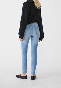 Vaqueros ajustados de color azul con detalles descoloridos y dos bolsillos traseros, combinados con un suéter negro de gran tamaño y zapatos planos negros.