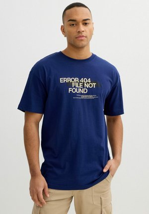 Hombre con una camiseta azul marino con el texto "ERROR 404 FILE NOT FOUND", pantalones cargo beige, de pie frente a un fondo claro y sencillo.