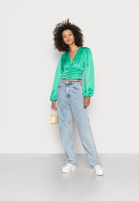 Top verde em satin com mangas bufantes, combinado com calças de ganga largas de um azul claro e ténis brancos com plataforma. A segurar uma pequena bolsa amarela.