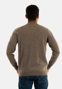 Man met kort krullend haar staat met zijn rug naar de camera, gekleed in een bruin sweater en een spijkerbroek, met zijn handen op zijn heupen.