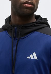 Κοντινό πλάνο ενός ατόμου που φοράει μπλε και μαύρο φούτερ με φερμουάρ και λευκό τριγωνικό λογότυπο στο στήθος.