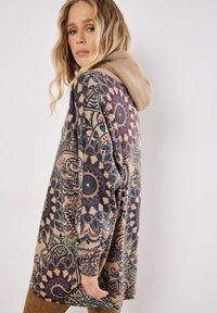 Sudadera con capucha de base beige, con intrincados patrones florales en azul y rojo, y un suave forro de felpa color marrón claro en la capucha.