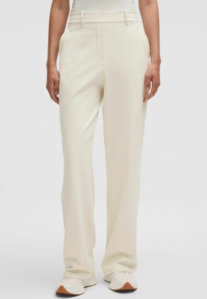 Kobieta w kremowych spodniach o prostych nogawkach i beżowych trampkach, stojąca na tle jednolitego białego tła.