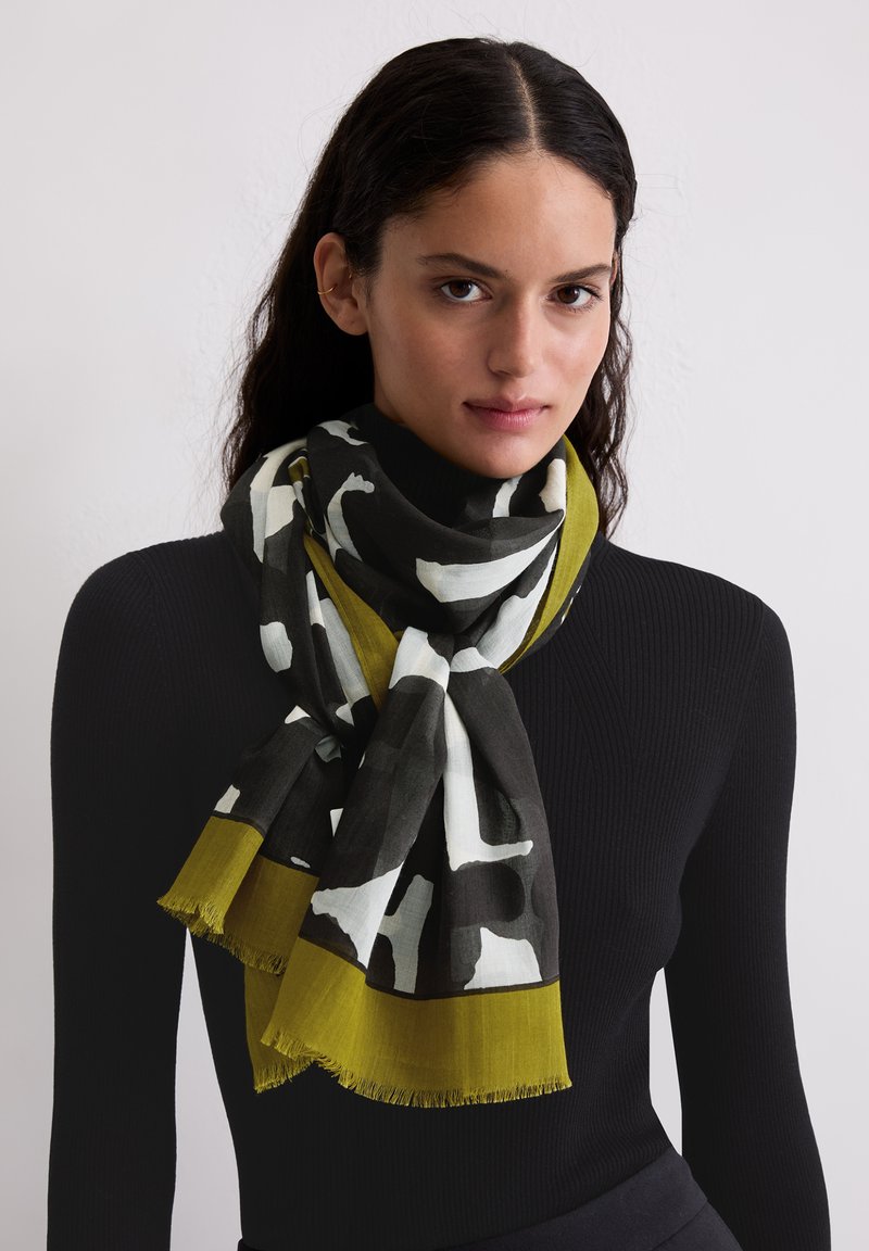 Sjaal met patroon in zwart, wit en olijfgroen, gemaakt van een lichtgewicht stof met franjes aan de randen. Gedrapeerd rond de hals.