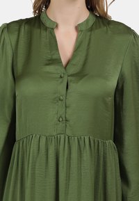 Blouse verte avec un col rond, fermeture à boutons et taille froncée. Texture lisse avec un léger éclat. Manches longues avec épaules bouffantes.