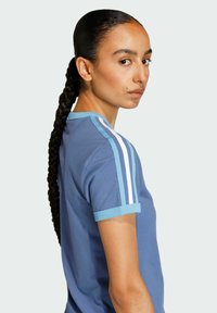 Niebieska koszulka z krótkim rękawem z jasnoniebieskim obszyciem i trzema białymi paskami na ramionach, wykonana z gładkiej tkaniny o luźnym kroju.