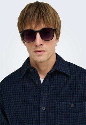 Jeune homme aux cheveux châtain clair portant des lunettes de soleil rondes foncées et une chemise à carreaux bleu foncé boutonnée sur un haut blanc.
