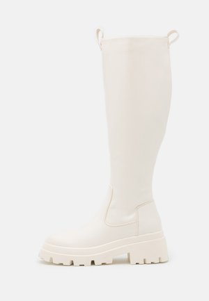 Botas de goma blancas hasta la rodilla con suela gruesa y paneles elásticos laterales. Cuenta con lengüetas en la parte superior para facilitar su colocación. Textura suave.