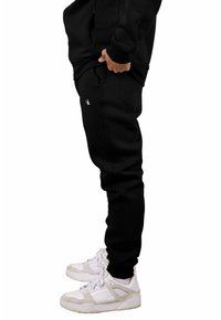 Pantalones deportivos negros de tela suave con un diseño ajustado, que cuentan con bolsillos laterales y un pequeño logo bordado en el muslo. Combinados con zapatillas deportivas blancas.