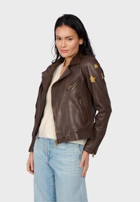 Veste de moto en cuir marron avec des accents d'étoile dorée sur les manches, dotée de plusieurs poches zippées et d'un design ajusté.