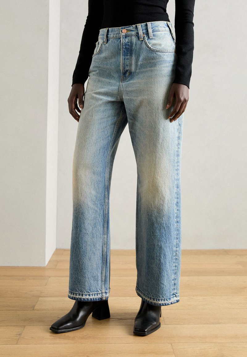 Jean large bleu clair en denim avec des zones décolorées, design à cinq poches et coutures contrastées. Porté avec des bottines noires à talons.