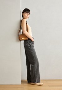 Beige ujjatlan felső, szürke bővülő farmernadrág, barna keresztpántos táska arany kiegészítőkkel, és világos színű laposlábú cipők. A modell a falnak dől.