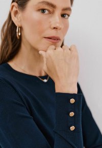 Granatowy sweter z rybami na mankietach, ozdobiony trzema złotymi guzikami. Tło jest neutralne. Ręka modelki spoczywa w zamyśleniu na brodzie.
