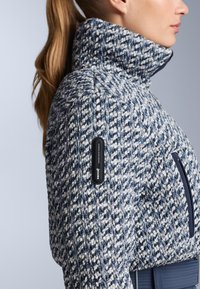Pletena jakna s visokim ovratnikom, plavo-bijelim teksturiranim uzorkom i bočnim džepom sa patentnim zatvaračem, s crnom RECCO etiketom.