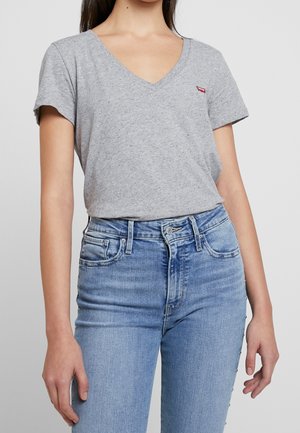 Femme portant un t-shirt à manches courtes en gris chiné avec un col en V, rentré dans un jean taille haute bleu avec des clous argentés sur les coutures extérieures.