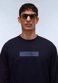 Μαύρο φούτερ με υφασμάτινο μπλε λογότυπο με το κείμενο «NAPAPIJRI geographic». Μοντέλο που φοράει γυαλιά ηλίου με ταρταρούγα σκελετό. Λείο ύφασμα.