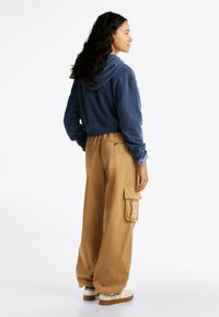 Felpa corta blu navy con cappuccio e cordino, abbinata a pantaloni cargo beige con tasca laterale e cuciture rosse, indossata con sneakers bianche.