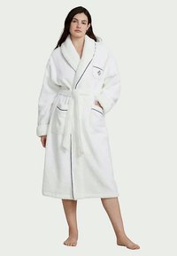 Valge pehme vannimantel, millel on šalli krae, seotud vöö, kaks taskut ja kontrastsed tumesinised aktsendid. Pehme tekstuur suurendab mugavust ja soojust.