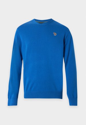 Jeune homme portant une chemise bleue à manches longues, un pantalon beige à jambes larges et des baskets blanches, debout sur un fond blanc uni.