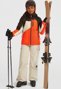 Tenue de ski comprenant une veste orange vif et marron, un pantalon beige, des chaussures de ski noires, des bâtons et des skis avec des motifs décoratifs.
