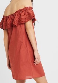 Trägerloses Kleid aus ziegelrotem Stoff, mit einem strukturierten Spitzenrüschchen am Ausschnitt und einer lockeren A-Linie-Silhouette.