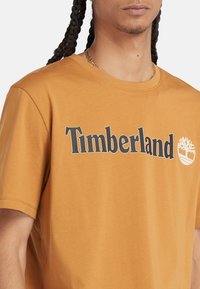 T-shirt in cotone arancione con logo "Timberland" in nero e una piccola grafica bianca di un albero. Maniche corte e design a girocollo.