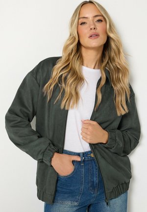 Donna bionda con capelli mossi indossa una giacca verde scuro con zip sopra una camicia bianca e jeans blu a vita alta, posando con una mano in tasca.