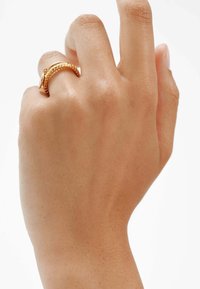 Anillo de oro con un diseño retorcido y banda texturizada, llevado en una mano con un tono de piel suave y natural.