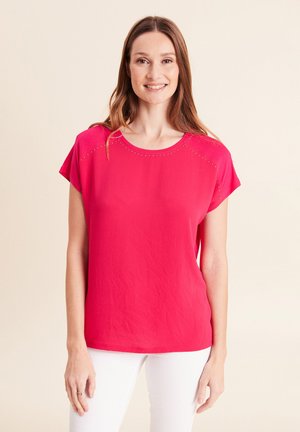 Donna sorridente, indossa una maglietta a maniche corte rosa acceso con piccoli dettagli a taglio vicino alla scollatura, abbinata a pantaloni bianchi, sfondo neutro.