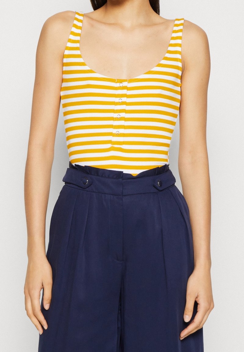 Femme portant un haut sans manches à rayures jaunes et blanches avec des boutons, associé à un pantalon plissé taille haute bleu marine.