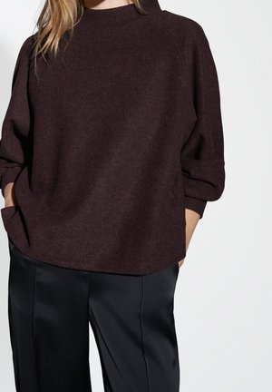 Osoba ubrana w ciemnobrązowy sweter z długim rękawem z rękami w kieszeniach oraz czarne, szerokie spodnie z satyny na jasnym, jednolitym tle.