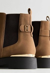 Bottines en suede marron avec un panneau en tissu côtelé de couleur sombre, des accents dorés et une semelle noire épaisse. Comprend un détail de sangle sur le côté.