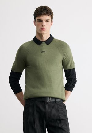 Junger Mann, der einen olivgrünen Kurzarmpullover mit schwarzem Kragen über schwarzem Langarmshirt trägt, schwarze Nadelstreifenhosen und einen schwarzen Gürtel.