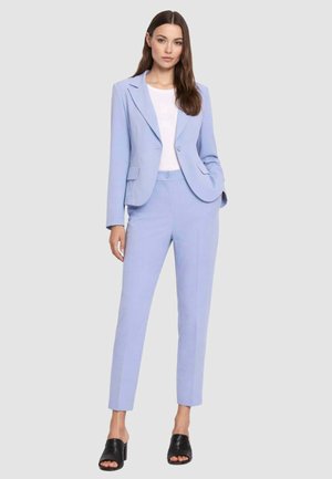 Donna che indossa un tailleur pantalone azzurro chiaro su misura con top bianco e mules nere con tacco e punta aperta, in piedi davanti a uno sfondo chiaro e uniforme.