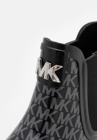 Bottine noire en caoutchouc à finition brillante avec un motif MK gris, agrémentée d'un logo argenté sur le côté.