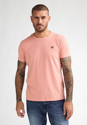 Hombre con pelo corto y tatuajes, vestido con una camisa rosa de manga corta y vaqueros azules, de pie frente a un fondo blanco liso.