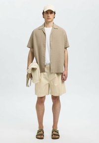Beige textuur kortegemouw shirt, witte T-shirt, beige shorts en olijfgroene schuifsandalen. Houdt beige doek in één hand.